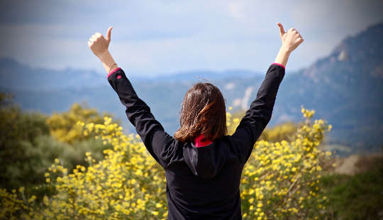 woman looking out over a valley with her two hands in the air with thumbs up