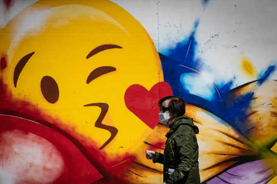 A man wearing a protective face mask and gloves walks past a large emoji face painted on the boarded-up windows of a store on Robson Street, in Vancouver, in May 2020. 