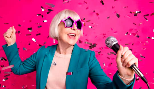 grey-haired woman wearing funky pink sunglasses singing holding a microphone