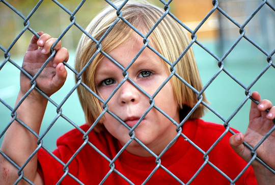 a young boy looking out from behind a chain-link fence