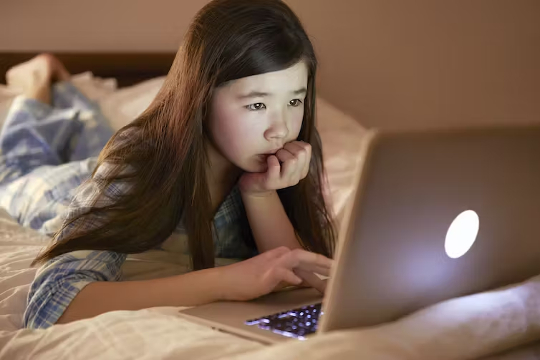 a young girl laying on her bed using a laptop under the eye of a webcam