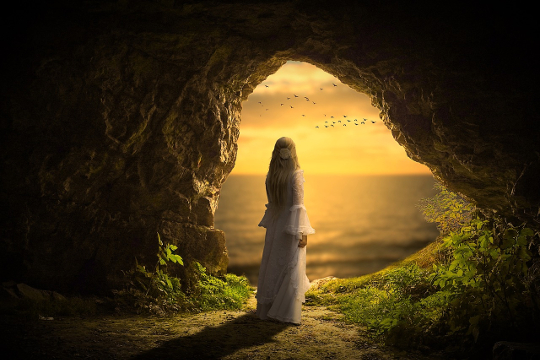 woman standing in a dark cave looking out into the bright sky