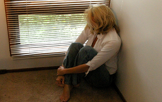 woman sitting on the floor looking out through the cracks of a venetian blinds of a window
