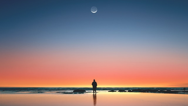 man standing on seashore facing a thin sliver of moon