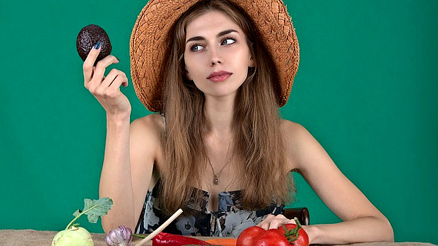 a woman with an array of fresh vegetables in front of her and holding up an avocado
